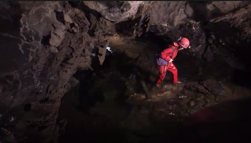Still from The Antibiotic Hunters Person in a red diving suit walking through a dark cave