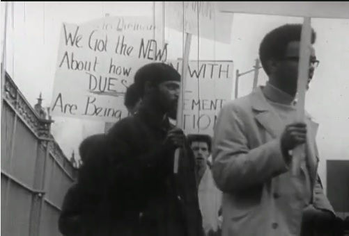 Two black men in a crowd carrying protest signs, one of which says We Got the News About how Dues Are Being (last word blocked from view)