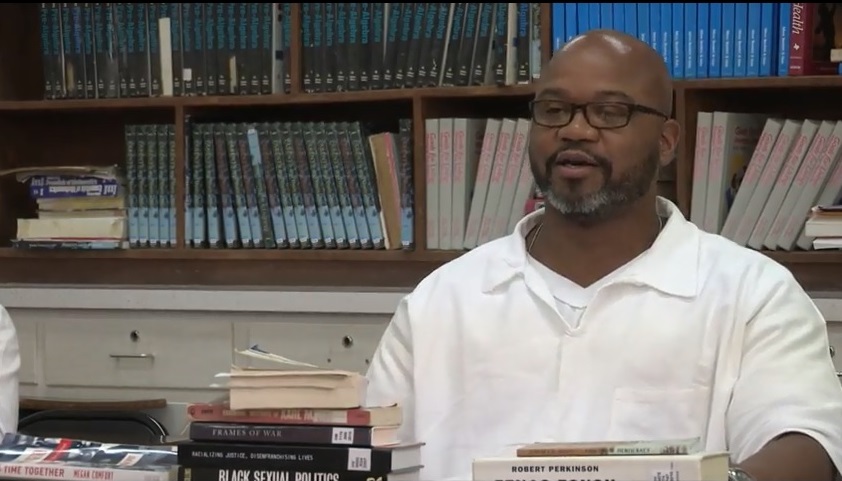 Middle-aged black man in glasses behind a desk covered in books, with a background of many books